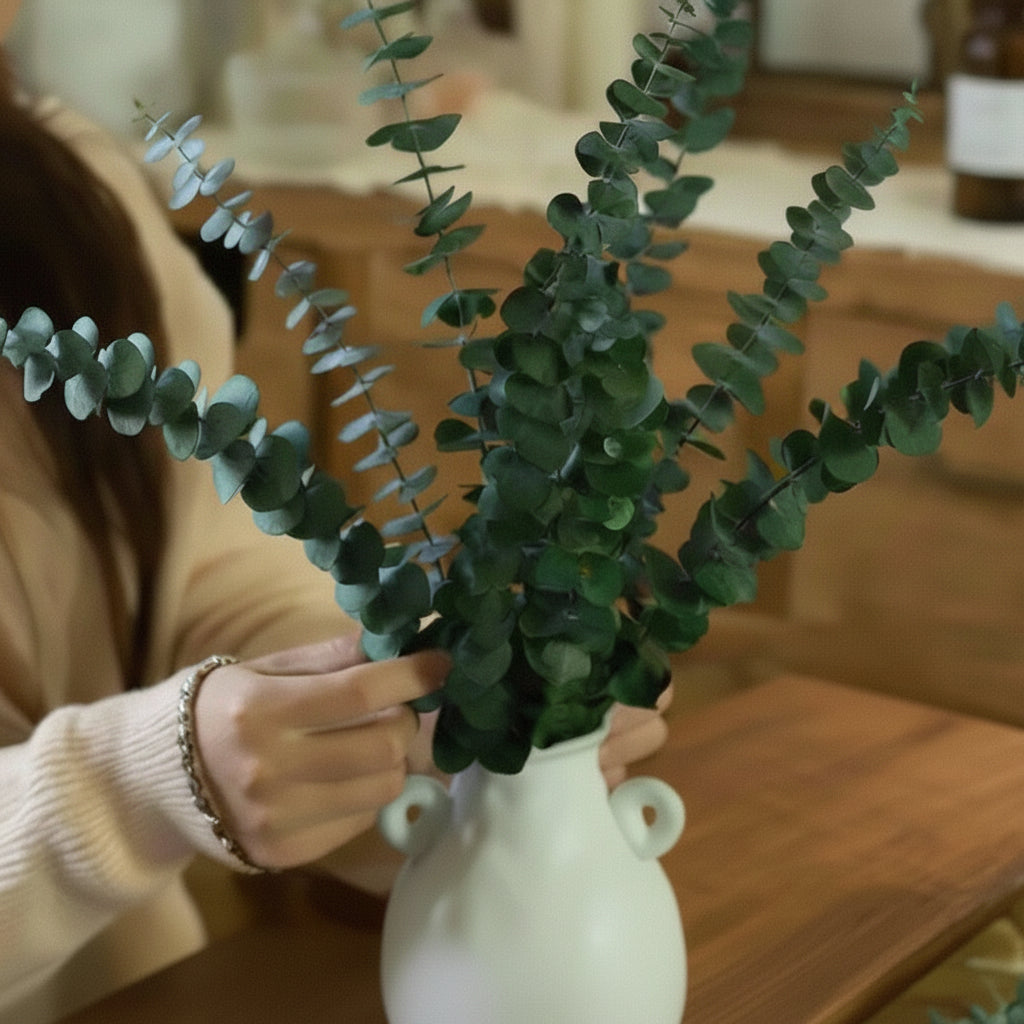 Person arranging eucalyptus greenery in a white vase on a wooden table